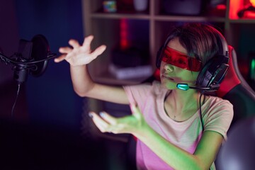 Wall Mural - Adorable hispanic girl streamer playing video game using virtual reality glasses at gaming room