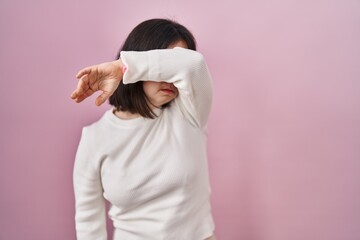 Wall Mural - Woman with down syndrome standing over pink background covering eyes with arm, looking serious and sad. sightless, hiding and rejection concept