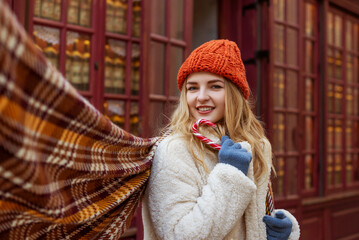 Wall Mural - Happy smiling woman wearing orange knitted beanie hat, gloves, checkered scarf, faux fur coat, holding candy cane, posing in street. Christmas, winter holidays concept. Copy, empty space for text