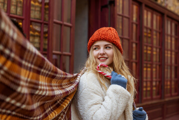 Wall Mural - Happy smiling woman wearing stylish knitted beanie hat, gloves, checkered scarf, faux fur coat, holding candy cane, posing in street. Christmas, winter holidays concept. Copy, empty space for text