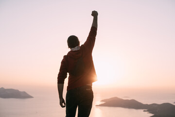 A young man meets the sunset, raising his hands in a victorious gesture on a mountain by the sea.