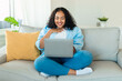 © Prostock-studio - Happy black oversize lady using laptop at home, reading news or making video call with friends, resting on sofa at home