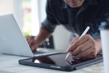 Close up of asian man using stylus pen on digital tablet pc and working on laptop computer on table. man illustrator, graphic designer working on mobile app
