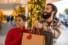Close Up Multiracial Couple Pointing To A Shop Window In A Mall With Christmas Trees And Lights. Happy African American Woman And Hispanic Man. Valentine's Day
