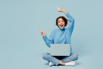 Full body young IT woman in knitted sweater hold use work on laptop pc computer point finger aside on area isolated on plain pastel light blue cyan background studio portrait People lifestyle concept.