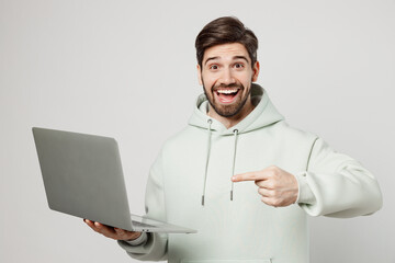 Young surprised amazed shocked caucasian man wear mint hoody hold use work point index finger on laptop pc computer isolated on plain solid white background studio portrait. People lifestyle concept.