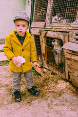 Little cute farm 2 years old boy, wearing autumn yellow coat and a cap feeding the chickens and the goat, cavy, rabbits in the countryside. Concept of friendship between children and animals