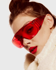 Wall Mural - Close-up portrait of young with well-kept skin, trendy makeup and red glasses posing in furry coat isolated over white background