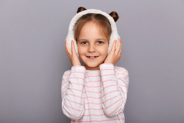 Wall Mural - Portrait of little cute female kid standing in fur headphones and looking at camera with smile, expressing positive emotions, wearing striped shirt, being in good mood.