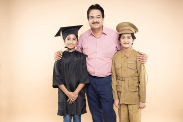 Wall Mural - Happy senior Indian man standing with girl child wearing convocation costume and boy kid wearing police uniform isolated over beige studio background.