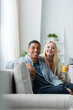 © LIGHTFIELD STUDIOS - blonde woman with glass of fresh orange juice sitting on couch near african american boyfriend and smiling at camera