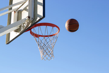 Wall Mural - Low angle shot of basketball flying into hoop outside on sunny day. Ball thrown into basket with white backboard against blue cloudless sky. Achievement, sports concept