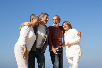 Two senior couples having fun outdoors on sunny day, standing against blue sky and laughing. Happy old friends enjoying leisure time together. Friendship, leisure, retirement concept