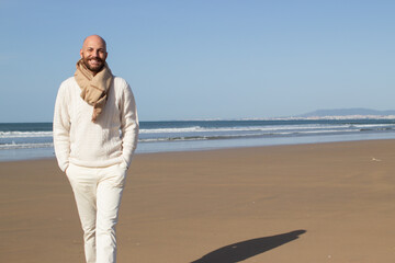 Wall Mural - Smiling handsome man walking alone on beach. Happy bald tourist in scarf holding hands in pockets of white jeans. Resting at sea concept