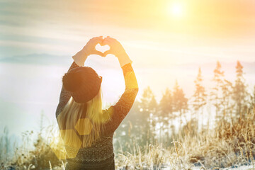 Happy traveler woman tourist stay on mountain top and making of love heart by hands in sunny frozen winter day.
