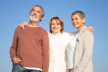 Portrait of three senior friends outdoors standing against blue sky background and smiling. Gray-haired bearded man and two middle-aged ladies enjoying time together on bright day. Friendship concept