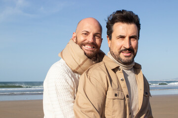 Wall Mural - Middle aged gay hugging partner from back. Smiling bald man in scarf and his bearded boyfriend in shirt looking at camera. Homosexuality concept