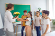 © Studio Romantic - Happy friendly children boys and girls congratulating elementary school teacher on professional holiday handing flowers to woman in gratitude for tutoring stand in classroom. Education for teens