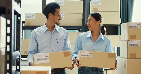 Wall Mural - Two happy Asian business success partnership man and woman wear uniform shirt carry cardboard box smiling looking at camera at warehouse. Good cooperation result, Startup small business concept.