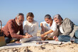 © KAMPUS - Group of people playing dominoes sitting on blanket at sandy shore. Bearded senior man making his move putting domino tile in row. Cropped shot. Recreation, leisure concept