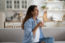Pleased Smiling Woman Resting On Sofa With Cup Of Tea Enjoying Alone Time At Home, Looking Aside With Dreamy Face Expression. Positive Female Starting Day With Morning Coffee. Free Time For Parent