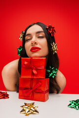 Canvas Print - Brunette model with shiny gift bows on hair posing near presents isolated on red