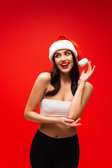 Canvas Print - Smiling brunette woman in top and santa hat posing isolated on red