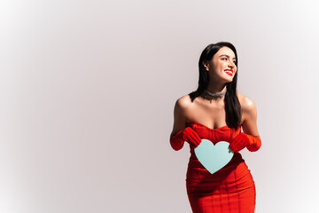 Canvas Print - Cheerful woman in red dress with naked shoulders holding paper heart isolated on grey