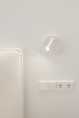 Bedside white LED wall lamp illuminating. Creative modern wall light source in bedroom. White European electrical socket outlet and switch. Electricity and illumination concept.