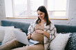 Smiling young expectant woman sitting on sofa and using laptop
