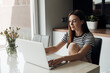 Pensive, smiling young brown long haired woman sitting in white kitchen, use laptop. Freelancer and self-employed worker