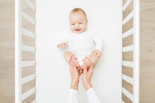 Young Mother Hands Holding And Massaging Infant Cute Small Bare Feet In White Crib At Home Room. Lovely Emotional Moment. Closeup. Parent Playing With Happy Adorable Smiling Baby Boy. Top Down View.