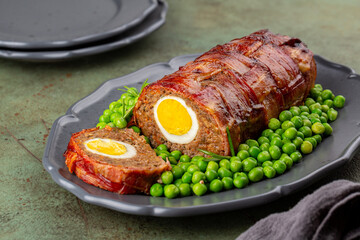 Wall Mural - Slice of baked meatloaf made of ground meat, onion, carrot, stuffed with hard-boiled eggs, wrapped with bacon net, served with green peas. German, Scandinavian and Belgian dish. Festive food.