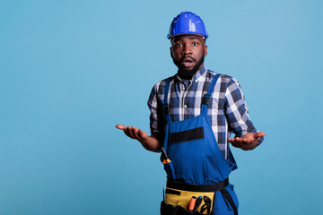Portrait of confused african american builder posing in work uniform on blue background, making uncertain facial expression. Man shrugging shoulders, studio shot I dont know concept.
