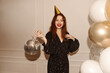 © Look! - Cheerful young caucasian girl celebrates her birthday in room with disco ball, air balloons. Red-haired beauty wears black dress and festive cap. Holiday concept