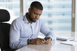 © fizkes - Serious young African business man reading, typing text message on smartphone, using online virtual app, chatting on Internet, reading, watching content, thinking, sitting at office work table