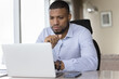 Serious focused young African business man using Internet app on laptop at office workplace, reading email letter, watching online presentation, business communication technology