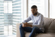© fizkes - Positive thoughtful young African business man using mobile phone for online communication, sitting on couch in modern home flat with big window glass, typing, chatting on Internet