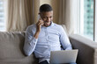 Happy millennial Black freelancer man, entrepreneur negotiating on business project on mobile phone, using laptop for online communication, sitting on couch, enjoying call, smiling, laughing