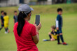 Mom standing and filming her son playing football in a school tournament on a sideline with a sunny day. Sport, outdoor active, lifestyle, happy family and soccer mom and soccer dad concept.