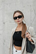 © alones - Urban fashion pretty woman with modern stylish black sunglasses in casual outfit with blazer, top and fashionable bag walks on the street near a grey concrete wall