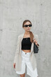 © alones - Trendy fashion hipster woman model with sunglasses in stylish casual outfit with blazer, top and shorts with bag stands near a gray wall on the street