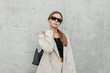 © alones - Stylish beautiful urban woman model with cool sunglasses in a fashionable beige blazer and a black top with a bag stands and poses near a gray concrete wall in the city