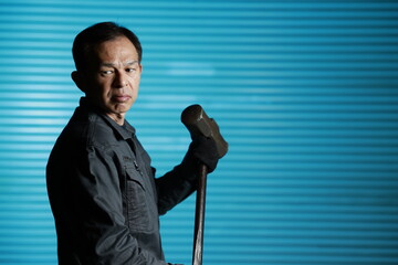 Craftsman in flayed work clothes holding a large metal hammer in front of the blue shutters of a town factory. Concept images of the essence of manufacturing and technical succession.