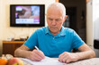 © caftor - Serious older man writing letter at table in living room