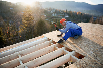 Wall Mural - Carpenter hammering nail into OSB panel on the roof top of future cottage at sunrise. Man worker building wooden frame house. Carpentry and construction concept.