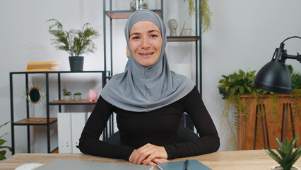 Muslim businesswoman in hijab working on laptop computer smiling friendly at camera and gesturing hello, hi, assalamu alaikum, greeting, welcoming with hospitable expression at office workplace desk