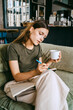 © Maskot - Young female entrepreneur writing in book holding coffee cup while sitting on sofa in studio