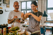 © Maskot - Happy female chef sprinkling chopped vegetable in pan while standing by colleague with diary at studio