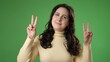 © Robert Peak - Portrait of happy young woman 20s smiling and giving peace or victory sign gesture with hands isolated on green screen background.